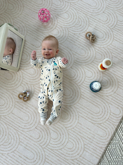 Baby lying on the Arch-pattern play mat, surrounded by a mirror, rattles and stacking toys
