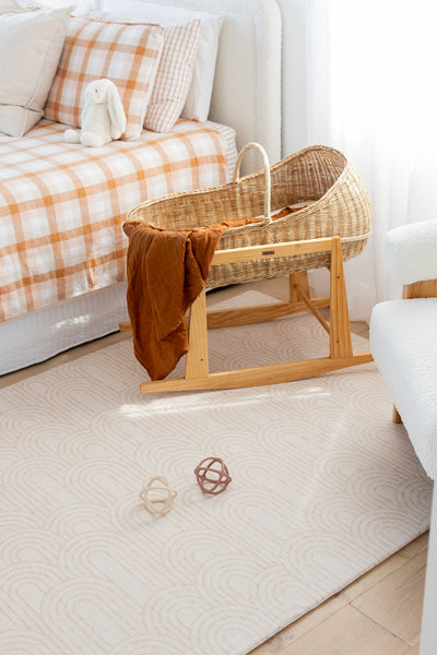 Arch-pattern baby play mat beneath a wicker bassinet on a wooden stand, with two toy balls